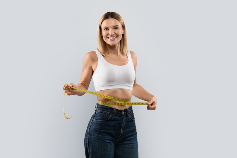 Happy pretty middle aged blonde woman wearing jeans and white top measuring her waist with measure tape, satisfied with diet or workout results, isolated on grey studio background, copy space